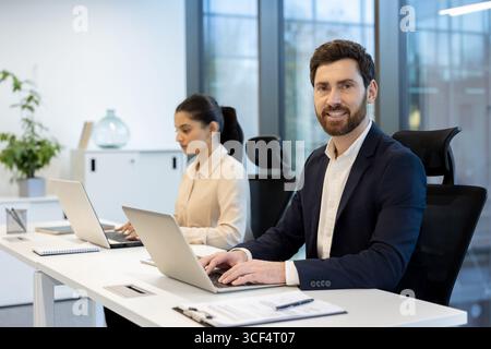 Coworkers are focused on tasks at laptops in a bright, modern office with natural light. The setting conveys productivity, professionalism, teamwork, and a collaborative work environment. Stock Photo