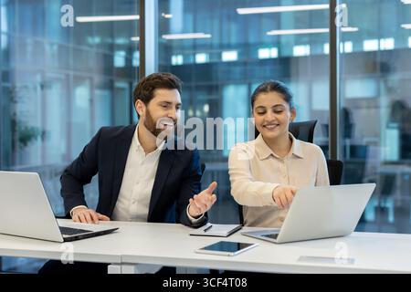 Two professional coworkers engaged in teamwork, using laptops while discussing ideas in a brightly lit office. Stock Photo