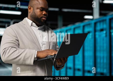 Software developer in server farm managing infrastructure systems, checking recovery plan. Data center IT specialist monitoring performance using notebook, making sure rigs are functioning optimally Stock Photo