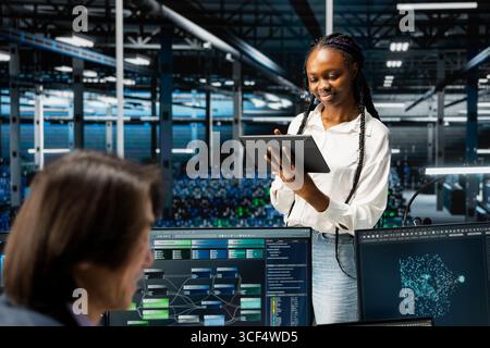 Smiling data center software developer using tablet, coding, managing systems for uptime and efficiency. Happy IT expert next to colleagues using programming languages in server hub Stock Photo