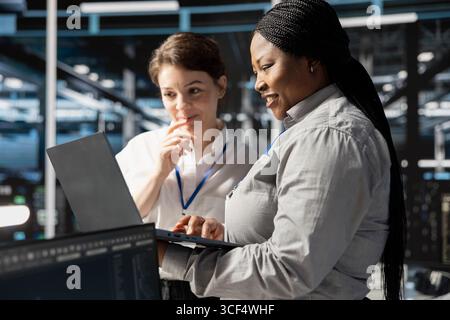 Smiling engineering team using laptop in data center, managing server virtualization, running scripts. Happy coworkers using programming language on notebook in server farm Stock Photo