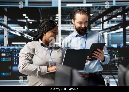 Happy system administrators in data center using devices, verifying firewall configurations. Jolly teamworking IT experts performing equipment vulnerability scans in server hub Stock Photo