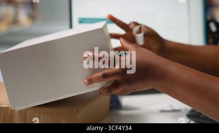 Black woman packing goods and applying shipping labels on boxes, creating awb tracking numbers on tags for easy distribution. Worker using adhesive airway bill stickers. Camera B. Stock Photo