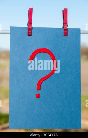 Red question mark with acrylic paint on blue drawing cardboard attached to a clothesline with two clothespins against a rural background. Stock Photo