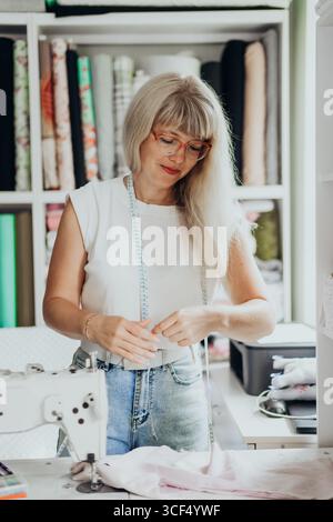 Smiling mature woman working as fashion designer in cozy studio surrounded by colorful fabric rolls and sewing tools measuring tape around neck pastel Stock Photo