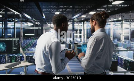 Data center team leader and worker reviewing performance dashboards of AI equipment rigs. Server room supervisor oversees programmer examining machine learning algorithms on screen, Stock Photo