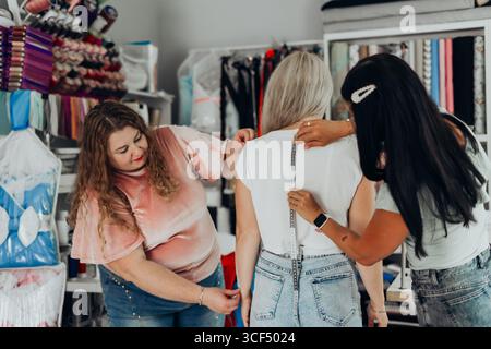 Two women measuring back length of woman with silver hair using tape measure in sewing studio filled with colorful threads fabrics and tools during Stock Photo