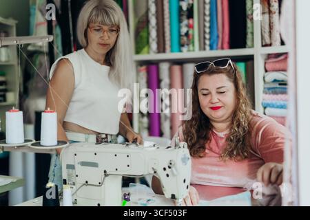 Two women collaborating in creative textile studio with sewing machine colorful fabric rolls discussing handmade garment design highlighting female Stock Photo