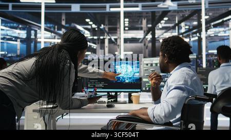 Data center engineer in wheelchair and colleague oversee automated tasks using node tree programming tool. Inclusive server farm team adjusts system settings based on AI analytics, camera A Stock Photo