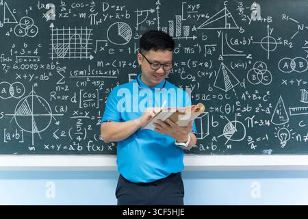 A mathematics teacher reading a book and writing notes while standing before a blackboard filled with equations, representing education, learning, aca Stock Photo