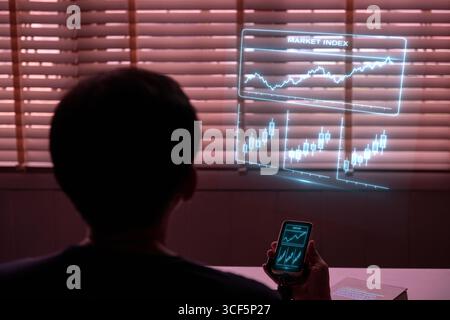 A person viewed from behind, using a phone to project a holographic stock chart. A concept for fintech, data visualization, investment strategy, virtu Stock Photo