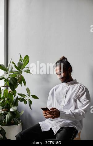 Vertical medium studio portrait of modern young Indian man wearing stylish white shirt sitting relaxed in office room reading something on smartphone Stock Photo