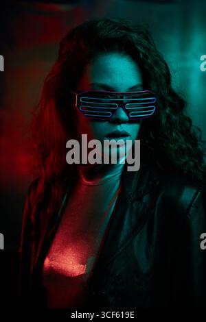 Vertical studio portrait of young Caucasian woman with long curly hair wearing futuristic eyeglasses and leather jacket looking away Stock Photo