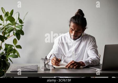 Horizontal medium portrait of young Indian man wearing fashionable white shirt and stylish accessories sitting in office room making notes Stock Photo