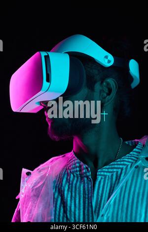 Vertical medium close-up studio portrait of stylish young Indian man wearing VR headset standing in neon light in darkness Stock Photo