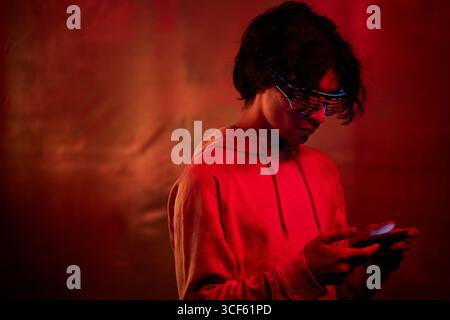 Portrait of handsome young Caucasian man with wavy hair wearing hoodie and neon eyewear standing in red and orange light playing game on smartphone Stock Photo