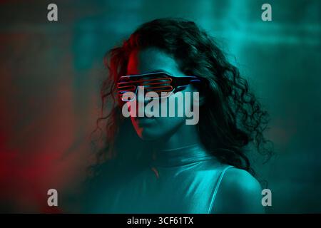 Studio portrait of young Caucasian woman with long curly hair wearing futuristic eyeglasses standing in neon light looking away Stock Photo