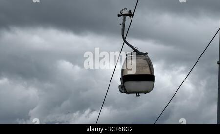 Cable car hanging in mid-air against a backdrop of dramatic, cloudy skies. This image captures the contrast between human engineering and the power of Stock Photo