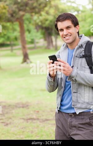 Man in his twenties standing in grassy park holding smartphone and wearing backpack, copy space Stock Photo