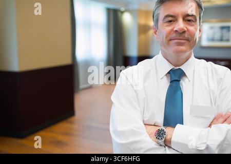 Middle-aged man in shirt and tie crossing arms in hotel corridor featuring wristwatch, copy space Stock Photo