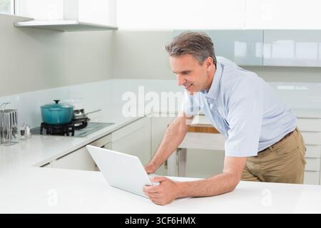 Middle-aged man leaning over countertop using white laptop beside teal pot in kitchen, copy space Stock Photo