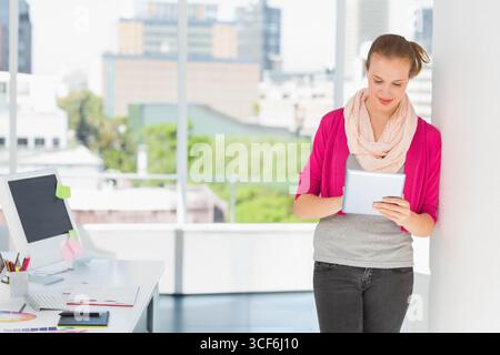 Woman reading tablet device at desk by office windows with computer monitor, copy space Stock Photo