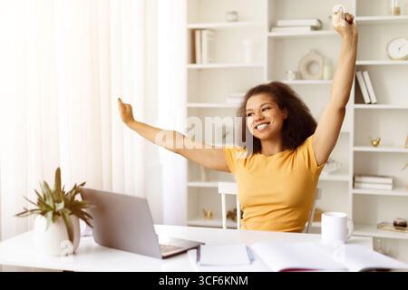 Cheerful African American woman stretching arms while sitting at desk with laptop Stock Photo