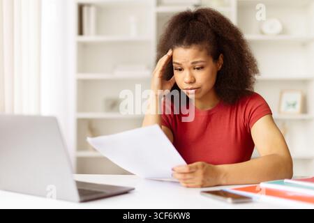 Young black woman looking stressed while reading documents at desk with laptop Stock Photo