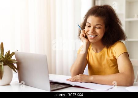 Smiling African American woman watching laptop screen and holding pen while studying Stock Photo