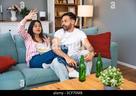 Young couple man and woman sitting at home on sofa taking a break after stressful work day watching sport game or movie on TV, eating popcorn and drin Stock Photo