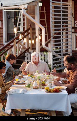 Group of multiethnic adults and senior Caucasian man sitting outdoors sharing meal and raising glasses during gathering, enjoying conversation around decorated table in backyard Stock Photo