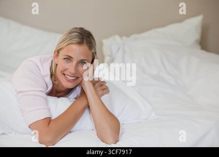 Woman lying on bed in bedroom wearing pink long sleeve shirt with white pillows, copy space Stock Photo