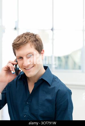 Man smiling and talking on smartphone while standing in office in front of large bright windows Stock Photo