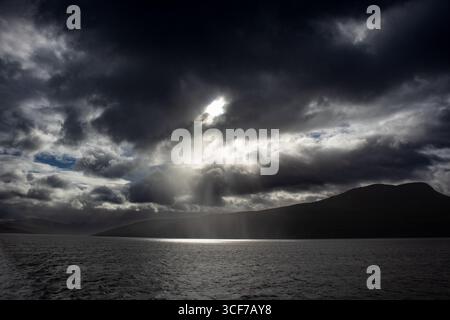 Sunlight breaking through heavy storm clouds over sea and mountains Stock Photo