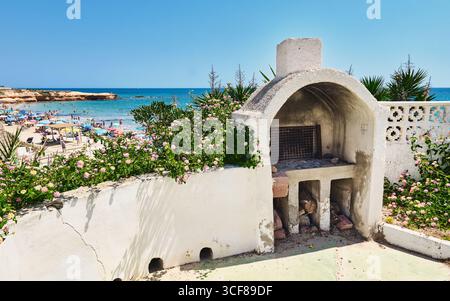 Weathered outdoor barbecue with cracked walls and flowers in the foreground, overlooking a crowded sandy beach with umbrellas and swimmers enjoying a Stock Photo