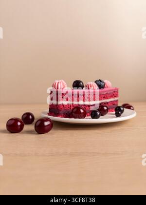 Close-up of red velvet cake with creamy filling, pink frosting swirls, and dark berries on a white plate. Set on a wooden surface with a beige backgro Stock Photo