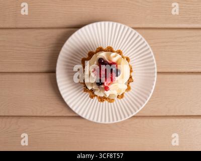 A centered tartlet topped with whipped cream and assorted berries, served on a white fluted plate over a wooden surface. Ideal for bakery promotions, Stock Photo