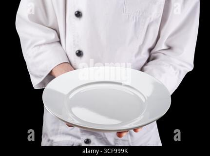 Chef in white uniform is holding an empty white round ceramic serving plate. Black background. Stock Photo