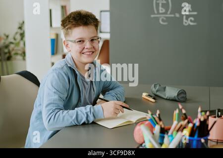 Portrait of Caucasian Boy Smiling While Writing in Notebook at Classroom Desk Stock Photo