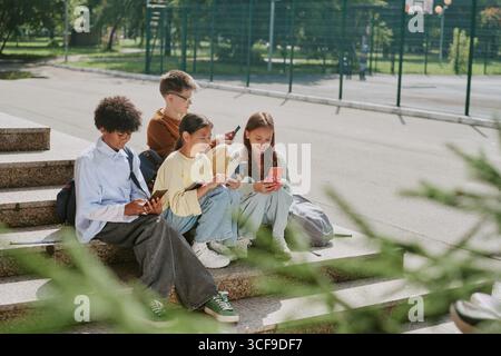 Group of Diverse Teenagers Sitting Outdoors Using Smartphones Together Stock Photo