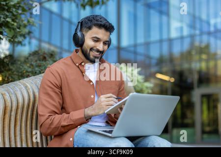 A man sits outdoors using a laptop and headphones, smiling and focused on tasks. Stock Photo