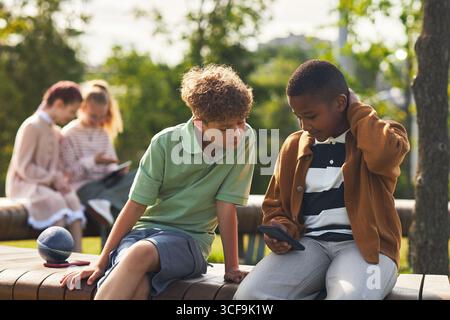 Two boys, one Caucasian and one Black, sitting outdoors on wooden bench, looking at smartphone together, two girls blurred in background using tablet Stock Photo