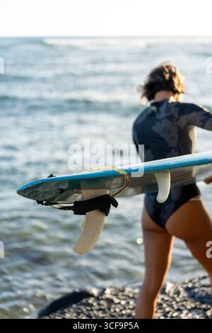 Blue surfboard with white fins in sharp focus as woman carries it out of frame—perfect shot emphasizing surf gear performance and style Stock Photo