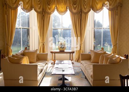 A luxurious living room features ornate gold curtains, plush seating, and large windows overlooking a serene garden view. Sunlight streams in, creatin Stock Photo