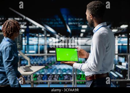 Data center programmer on industrial platform using green screen tablet, solving optimization tasks around the server farm. Male worker visualizing system performance using mockup screen. Stock Photo