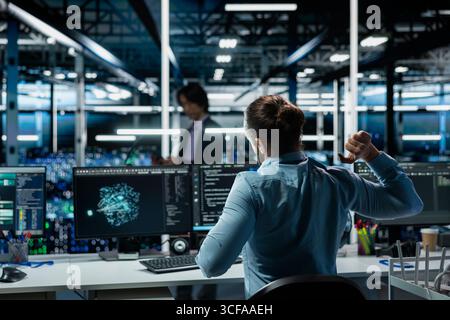 Data center engineer stretching at workstation to relieve back fatigue while using AI LLM assistant to perform maintenance. Server hub worker relaxing stiff back after long hours at computer Stock Photo