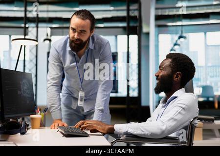 Manager supervising technician in wheelchair using AI on computer to optimize performance and forecast issues. Team leader and employee with disability in inclusive office train deep learning models Stock Photo