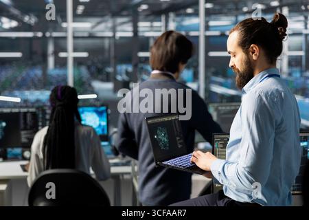 Engineer using laptop in data center with gear managing AI training datasets. IT expert using notebook, overseeing deep learning gear in server hub supporting scalable infrastructure for AI apps Stock Photo