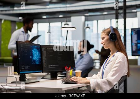 Smiling junior dev in tech startup utilizing AI to optimize performance while listening music. Upbeat female engineer wearing headphones writing code in office, training machine learning models Stock Photo