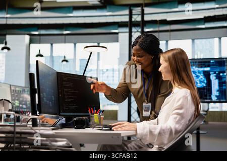 Friendly senior engineer mentoring jolly junior dev at workstation in office, reviewing AI system metrics. Happy african american manager guiding intern through code reviewing using machine learning Stock Photo